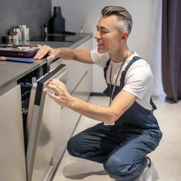 A man in overalls repairs a dishwasher, showcasing his skills as a kitchen installer in a modern kitchen setting.